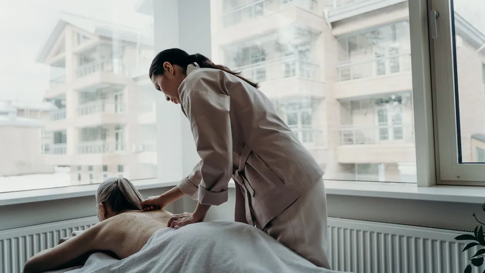 身穿米色制服的按摩师在明亮的窗边为盖着床单的顾客按摩背部